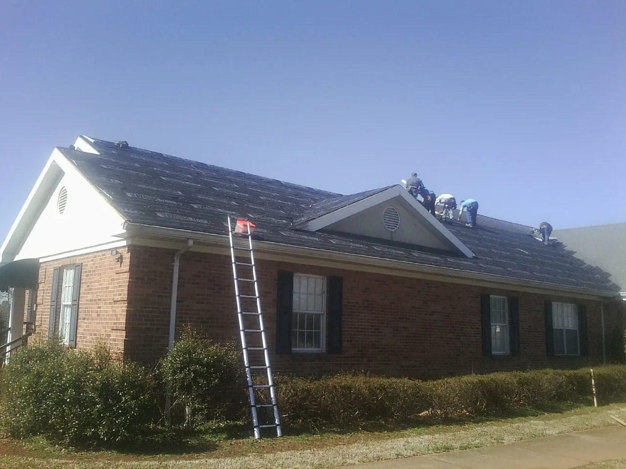 Crew tearing shingles off a brick ranch roof with a ladder set — Emergency Roof Repair work by our crew in Reva