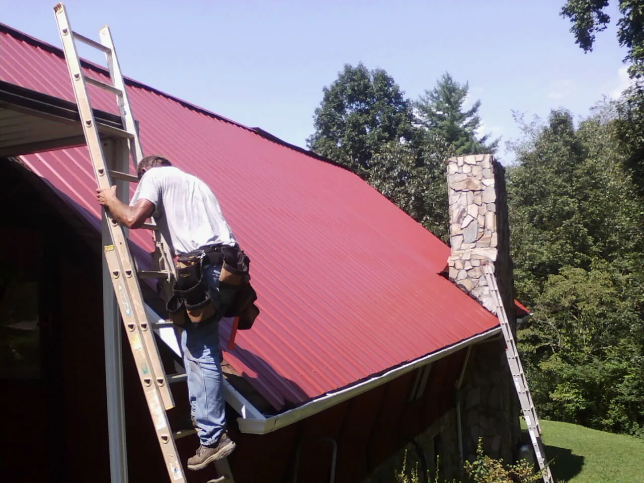 Roofer on a ladder beside a red metal roof and stone chimney — Soffit & Fascia Repair by Roofing Brothers in Reva